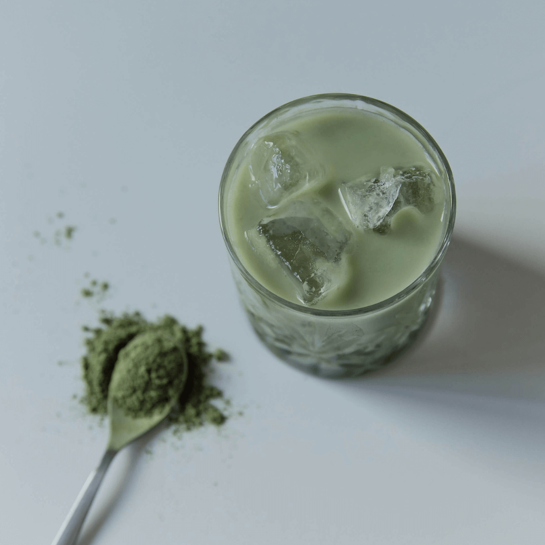 Ein stilvolles Riffle-Glas gefüllt mit einem erfrischenden Matcha Latte, perfekt für moderne Trinkerlebnisse. Neben dem Glas liegt ein Löffel mit Matcha-Pulver, um das Getränk individuell zu verfeinern.